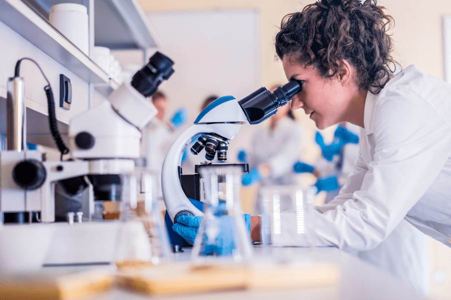 Wissenschaftlerin im weißen Kittel untersucht Proben durch ein Mikroskop – Qualitätskontrolle und Laboranalysen für die sichere Herstellung von Nahrungsergänzungsmitteln