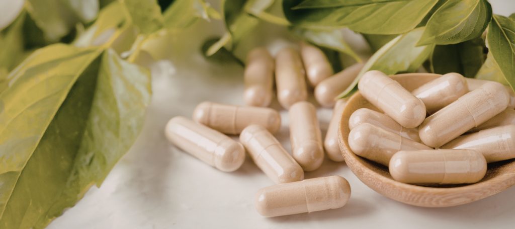 Beige Kapseln in einer kleinen Holzschale, umgeben von grünen Blättern auf hellem Untergrund